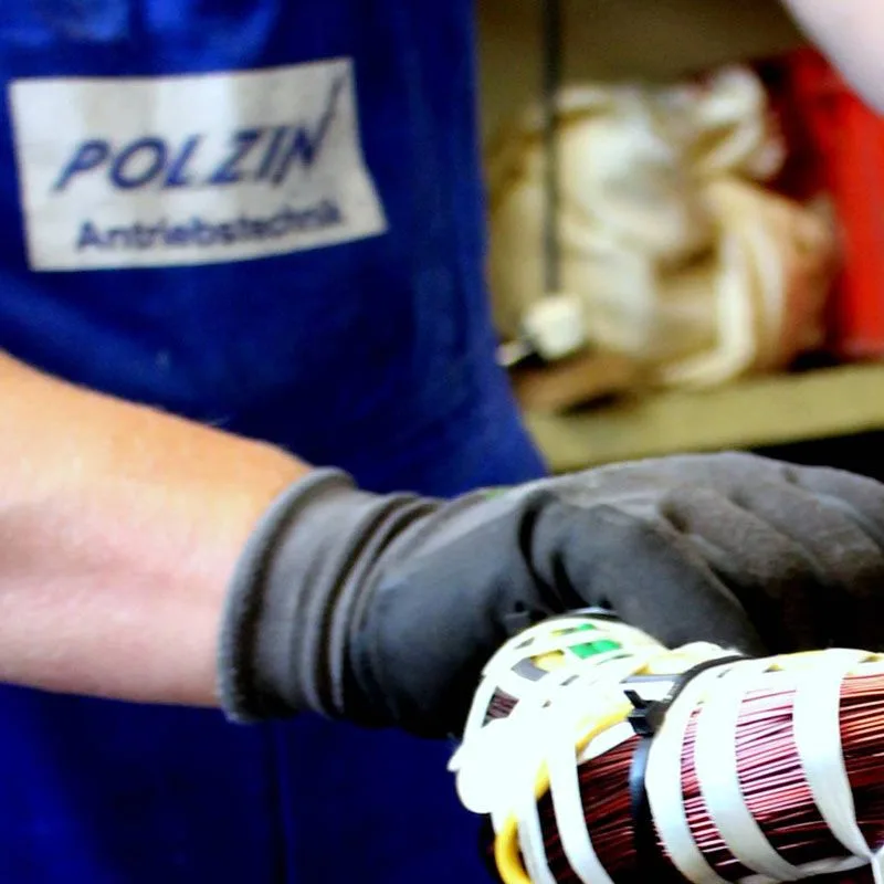 Hand im Arbeitshandschuh hält eine bewickelte Spule, blaue Schürze mit Aufdruck POLZIN Antriebstechnik